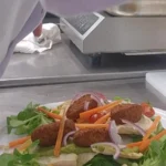 Assiette de keftas végétales sur mâche et crudités, tomates cerises, bâtonnets de carotte et oignon rouge, posée sur le plan de travail inox