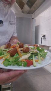 Assiette de keftas végétales sur lit de mâche et crudités, tenue devant le passe d'une cuisine de collectivité.