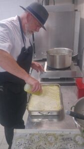 Un chef arrose de béchamel un bac de lasagnes en cours de montage, en cuisine de collectivité.
