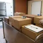 Quai de réception d'une cuisine collective : colis sur un chariot inox, sonde thermométrique piquée dans un carton, fiche de réception, camion frigorifique au fond
